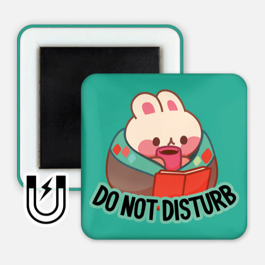 A square magnet featuring a cute bunny wrapped in a cozy blanket, holding a red book and a pink mug. The background is teal, and the text "DO NOT DISTURB" is written in bold black letters beneath the bunny. A small magnet icon is displayed in the corner, indicating the product type.