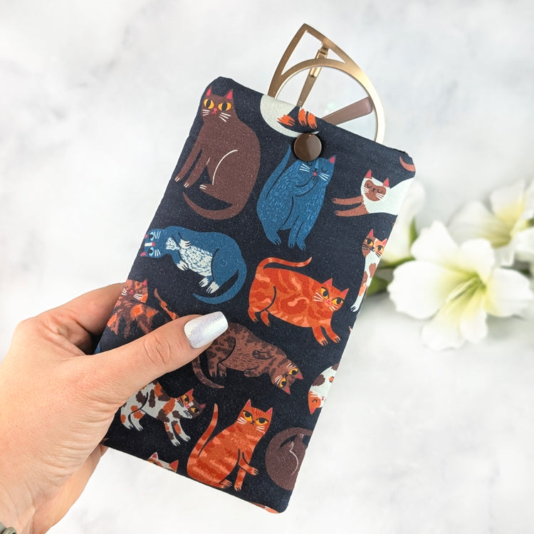 Hand holding a padded eyeglass sleeve featuring colorful illustrated cats on a navy background, with gold cat-eye glasses tucked inside and a brown snap closure.