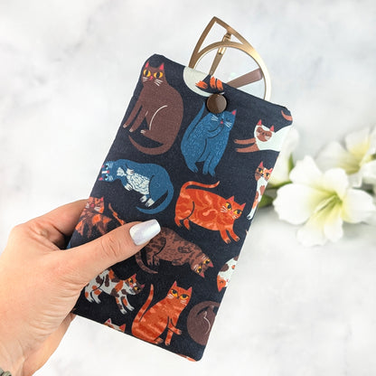 Hand holding a padded eyeglass sleeve featuring colorful illustrated cats on a navy background, with gold cat-eye glasses tucked inside and a brown snap closure.