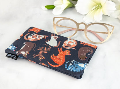 Flat-lay of a handmade padded eyeglass case with multicolored cats on a dark background, shown with cat-eye glasses beside it and white flowers for decor.