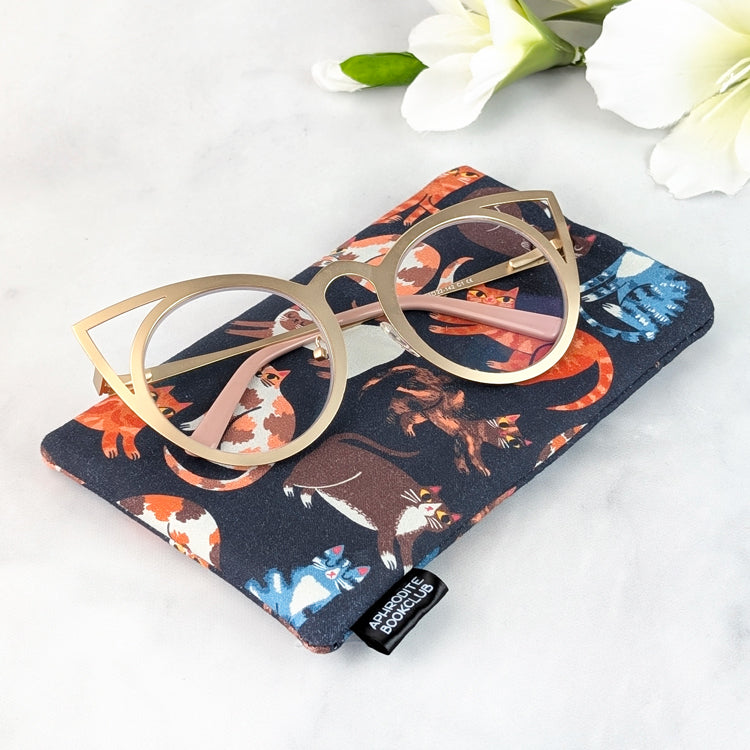 Close-up of a soft padded glasses sleeve with playful cat illustrations, displayed with cat-eye glasses on top and Aphrodite Bookclub fabric tag visible.