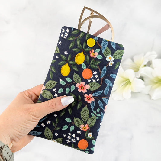 Hand holding a padded eyeglass sleeve with a vibrant citrus and floral pattern on a navy background, featuring a yellow snap closure and gold cat-eye glasses.
