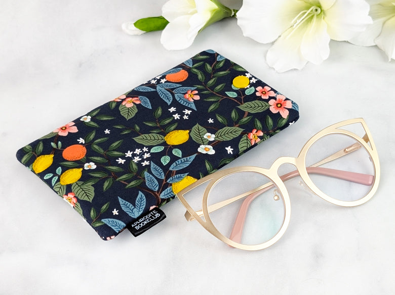 Flat-lay of a handmade padded eyeglass case with a citrus floral design on dark fabric, shown beside gold cat-eye glasses and white flowers in the background.