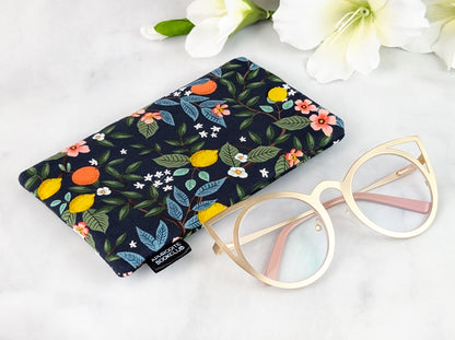 Flat-lay of a handmade padded eyeglass case with a citrus floral design on dark fabric, shown beside gold cat-eye glasses and white flowers in the background.