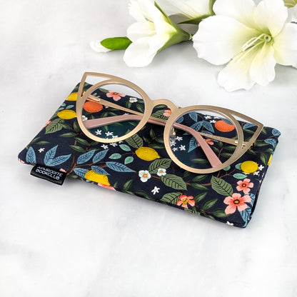 Close-up of a soft padded eyeglass sleeve with colorful lemon and orange branch pattern, displayed with cat-eye glasses on top and Aphrodite Bookclub tag visible.