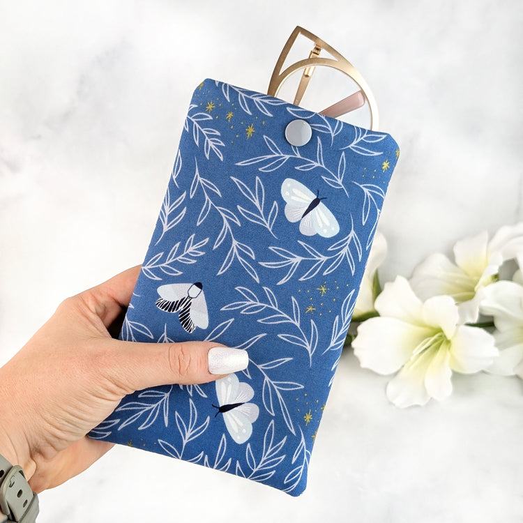 Hand holding a padded eyeglass sleeve in blue with white moth and leaf pattern, featuring a gray snap closure and gold cat-eye glasses peeking out.