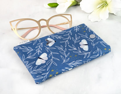 Flat-lay of a handmade padded eyeglass case with moth and foliage design on blue fabric, shown beside gold cat-eye glasses and white flowers.