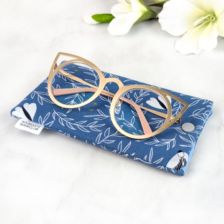 Close-up of a blue fabric eyeglass sleeve with silver moth print, displayed with gold cat-eye glasses on top and Aphrodite Bookclub tag visible.