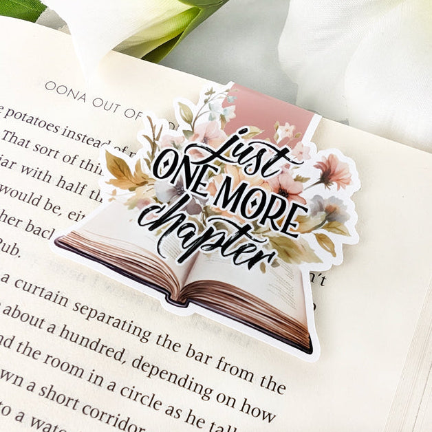 Magnetic bookmark featuring intricate floral designs in pastel colors and "Just one more chapter" written in italic, laying on an open book.