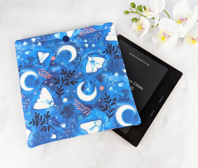 padded kindle oasis sleeve with snap. fabric showcases whimsical illustrations of moths, florals and moons on a blue background.