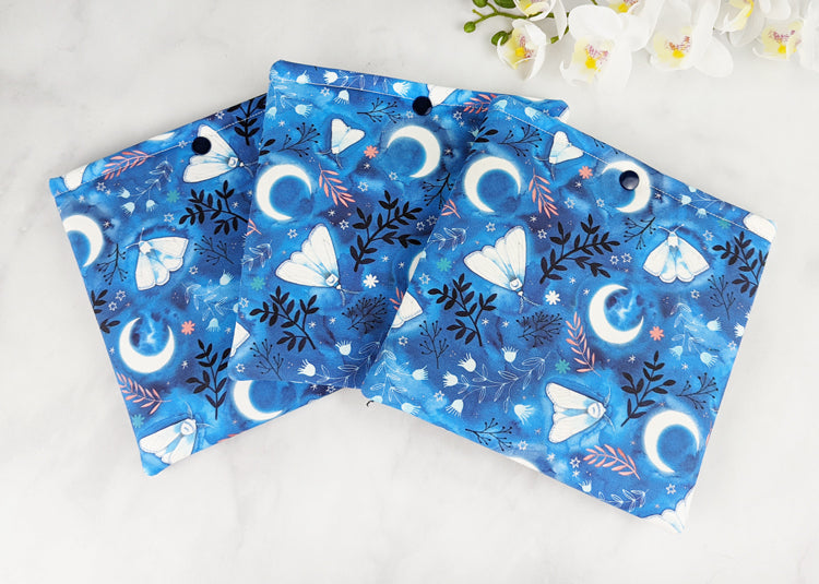 three padded kindle oasis sleeves with snap. fabric showcases whimsical illustrations of moths, florals and moons on a blue background.