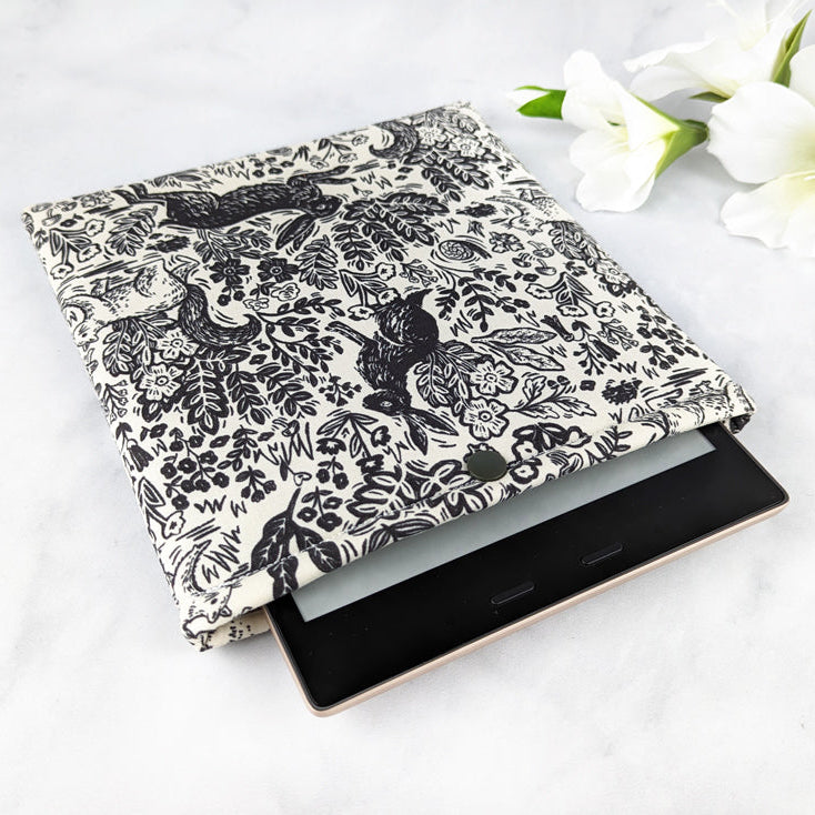 padded kindle oasis sleeve with snap. fabric showcases monochromatic vintage style line art featuring woodland elements like rabbits, squirrels and foliage on a off-white background.