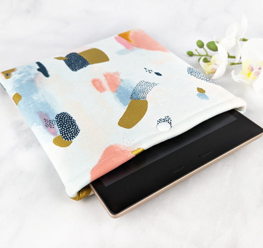 padded kindle oasis sleeve with snap. fabric showcases paint brush strokes in shades of navy blue, gold, pink and gray on a white background.