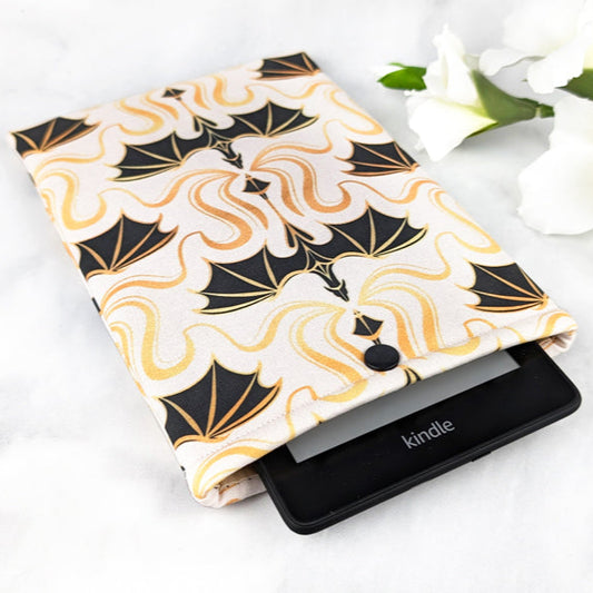 padded kindle paperwhite sleeve with snap. fabric showcases simple illustrations of flying dragons in a damask style in shades of black, gold and white.