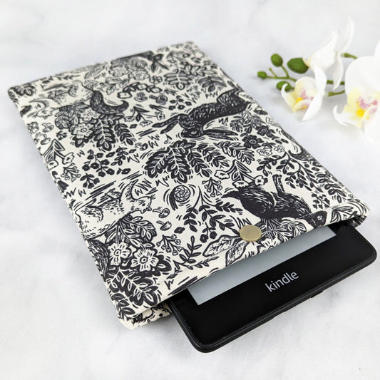 Kindle Paperwhite sleeve with a black and white woodland print featuring rabbits, birds, flowers, and foliage.