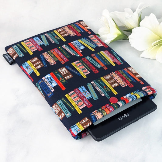 Handmade e-reader sleeve made from navy cotton fabric featuring colorful rows of illustrated classic book spines, including titles like Heidi and Gulliver’s Travels. The padded sleeve is shown holding a Kindle Paperwhite, displayed on a white marble background with white flowers for decoration.