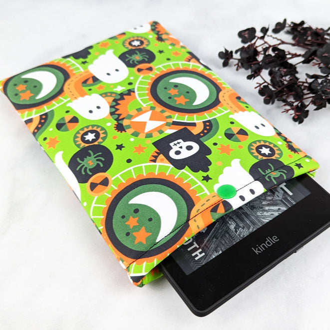 Bright green Kindle sleeve with a playful Halloween print featuring ghosts, skulls, moons, stars, spiders, and candy shapes in orange, black, and white.