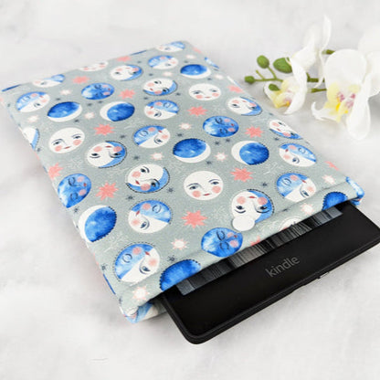 padded kindle paperwhite sleeve with snap. fabric features blue watercolor-style moon phases with faces on a light gray background.