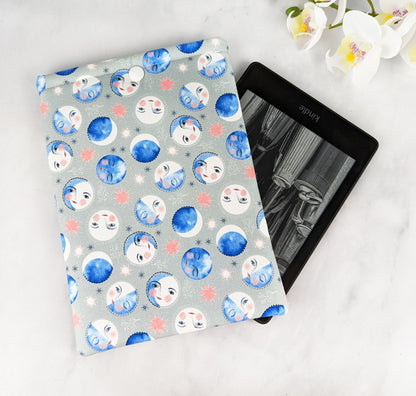 padded kindle paperwhite sleeve with snap. fabric features blue watercolor-style moon phases with faces on a light gray background.