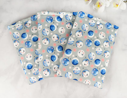 three padded kindle paperwhite sleeves with snap. fabric features blue watercolor-style moon phases with faces on a light gray background.