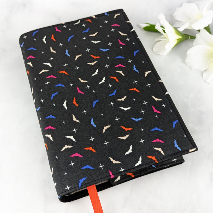 large adjustable book cover, fabric features mini bat shapes in shade of orange, blue, pink and white, on a black background.