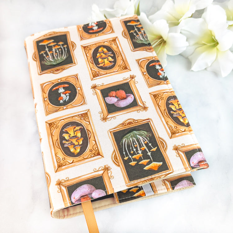 Fabric book cover featuring a whimsical mushroom gallery print with golden ornate frames on a cream background, showcasing various fungi illustrations in earthy orange, yellow, and plum tones.
