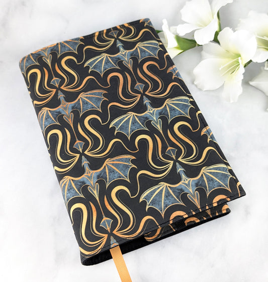 Fabric book cover with a bold pattern of gold and gray dragons on a black background, displayed with white flowers on a white surface.