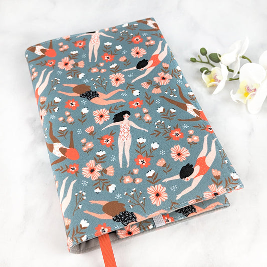 Fabric book cover with a whimsical design of illustrated women swimming and floating among coral and white flowers on a muted blue background, finished with an orange ribbon bookmark.