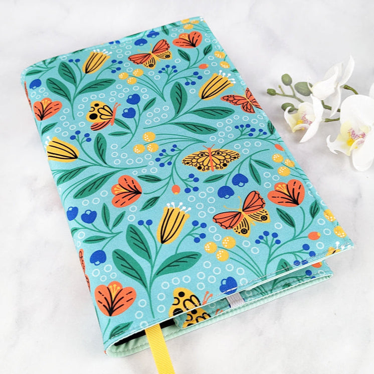 large adjustable fabric book cover, fabric features orange and yellow butterflies and flowers on a turquoise background.