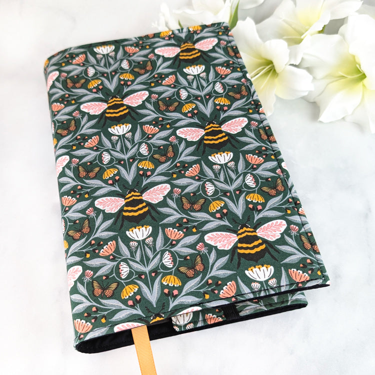 Dark green book cover with a detailed bee and butterfly pattern surrounded by pink, white, and yellow wildflowers on leafy vines.