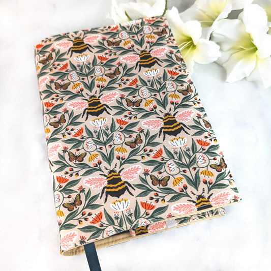 Beige book cover with a charming bee and butterfly pattern surrounded by pink, white, and yellow wildflowers on leafy green vines.