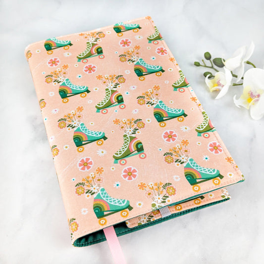 Fabric book cover with a playful retro design featuring colorful roller skates filled with flowers and scattered daisies on a soft peach background, finished with a light pink ribbon bookmark.