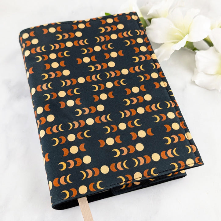 adjustable fabric book cover, fabric pattern features moon phases in beige and orange shades on a dark navy background.