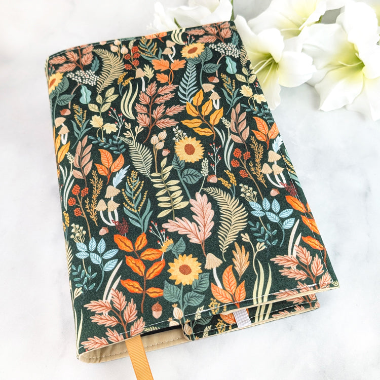 Dark green book cover featuring an intricate woodland pattern with autumn leaves, sunflowers, mushrooms, and ferns in warm orange, gold, and muted blue tones.