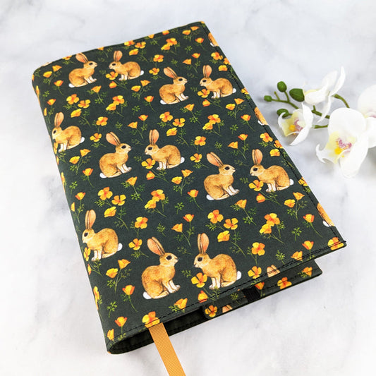 Fabric book cover with a whimsical print of golden brown rabbits and scattered orange wildflowers on a deep green background, finished with a golden yellow ribbon bookmark.