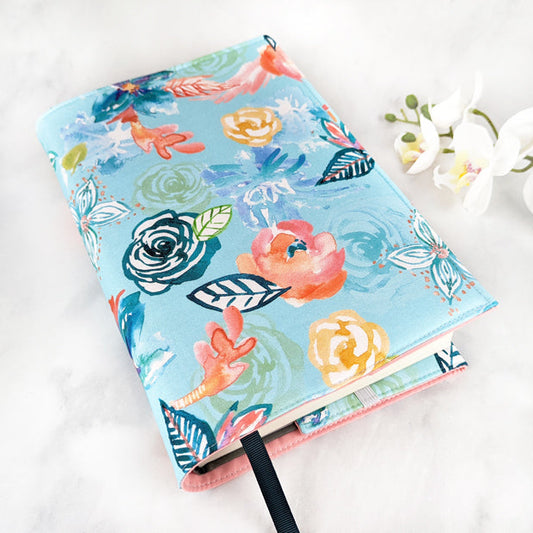 Fabric book cover with a vibrant floral watercolor design in shades of coral, yellow, navy, and green on a light blue background, finished with a navy ribbon bookmark.