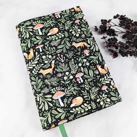 Fabric book cover with a woodland-themed design featuring orange foxes, red mushrooms, and lush green foliage on a black background, finished with a green ribbon bookmark.