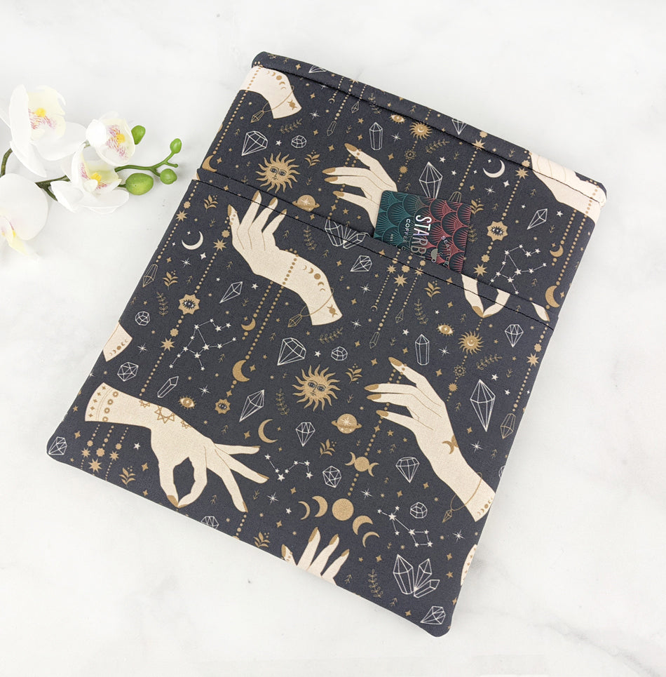Flat lay of a fabric book sleeve with a mystical design featuring illustrated hands, moons, suns, stars, constellations, and crystals in gold and cream on a dark navy background.