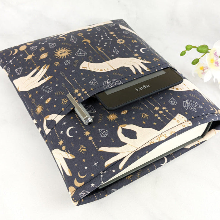 Fabric book sleeve with celestial and mystical hand design in gold and cream on a dark navy background, shown holding a Kindle, hardcover book, and pen. 