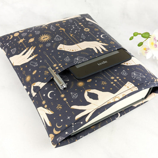 Fabric book sleeve with celestial and mystical hand design in gold and cream on a dark navy background, shown holding a Kindle, hardcover book, and pen. 