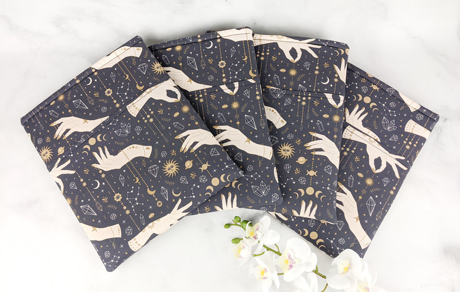 Four fabric book sleeves displayed in a fanned arrangement, each featuring a celestial and mystical design with cream-colored hands, gold moons, stars, suns, and crystals on a dark navy background. 