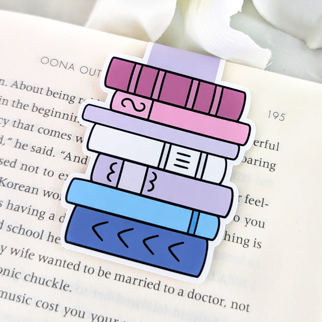 Magnetic bookmark shaped like a stack of books in bigender pride flag colors—shades of lavender, white, pink, and blue—attached to the top of an open book. 
