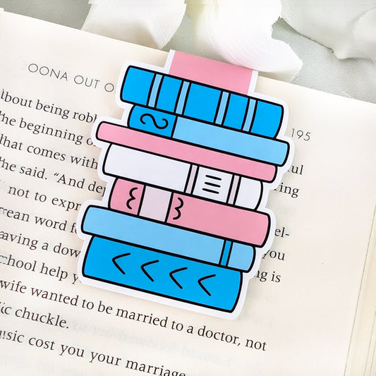 Magnetic bookmark in the shape of a pastel book stack in blue, pink, and white, clipped to the top of an open book. The colors represent the transgender pride flag. 