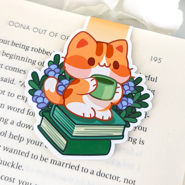 Magnetic bookmark featuring a cute orange cat holding a green mug while sitting on stacked books surrounded by purple flowers.