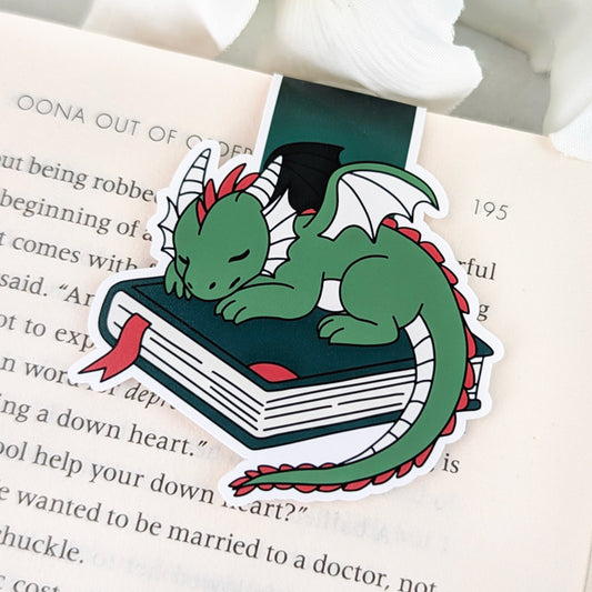 Magnetic bookmark featuring a cute green dragon with red accents sleeping on a closed book with a red ribbon bookmark.
