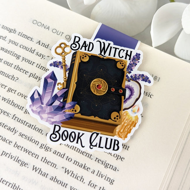 Magnetic bookmark featuring a spellbook surrounded by crystals, lavender, a candle, and the phrase 'Bad Witch Book Club,' clipped to an open book.