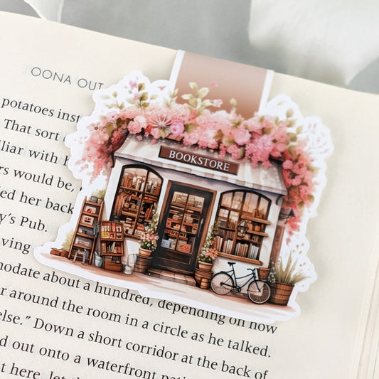 A colorful magnetic bookmark featuring a charming bookstore with a sign that reads "BOOKSTORE." The bookstore is adorned with pink flowers and has a bicycle parked outside.