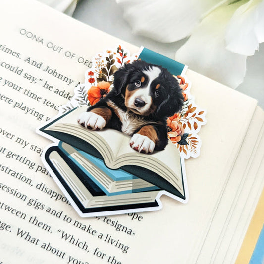 Colorful magnetic bookmark featuring a Bernese Mountain Dog puppy peeking out from behind a stack of books. The bookmark has a whimsical, hand-drawn
style with floral accents.