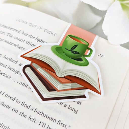 Magnetic bookmark with green tea cup on stack of books, clipped to an open book with white flowers nearby.