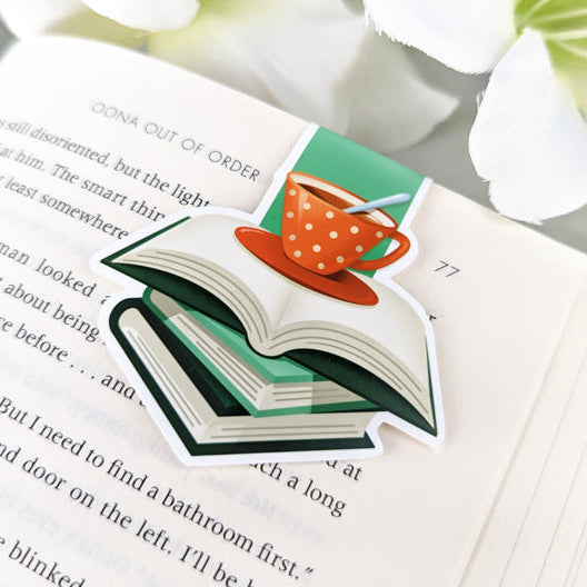 magnetic bookmark featuring an illustration of an open book stacked on top of two closed books with green covers. Above the books sits a red polka-dot teacup on a saucer with a spoon.